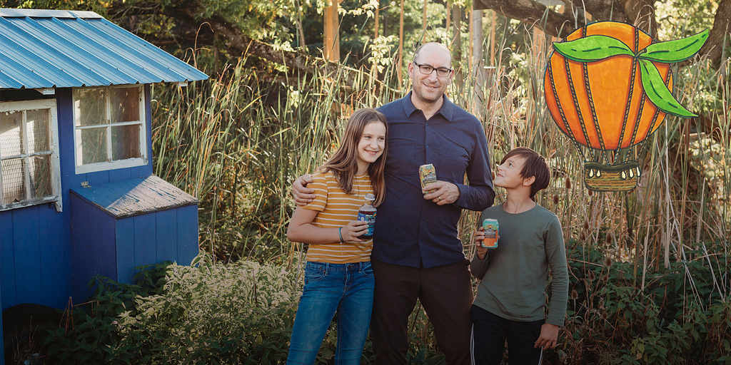 founder jeff weaber with his two children holding a can of kombucha with a floating orange hot air balloon illustration overlaid on the image