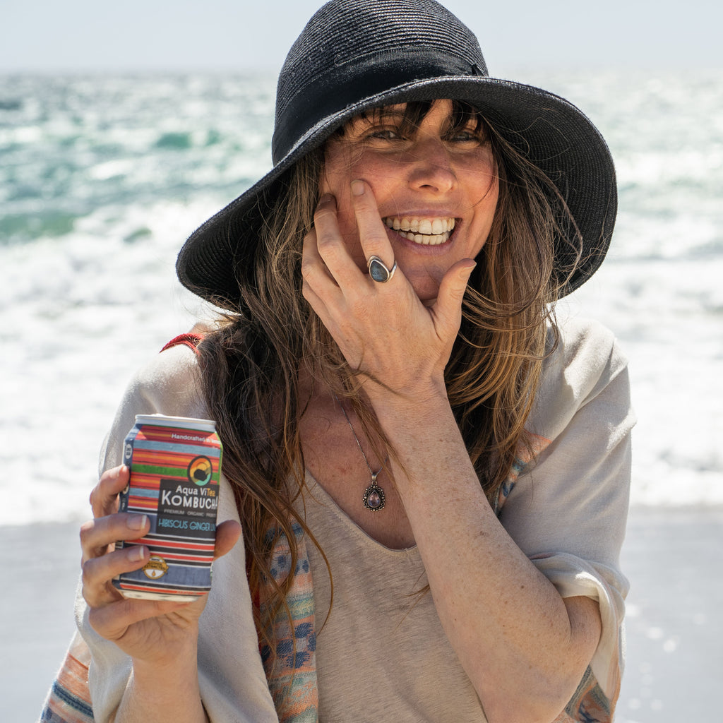 woman smiling on the beach near the water's edge drinking a can of hibiscus ginger lime aquavitea kombucha